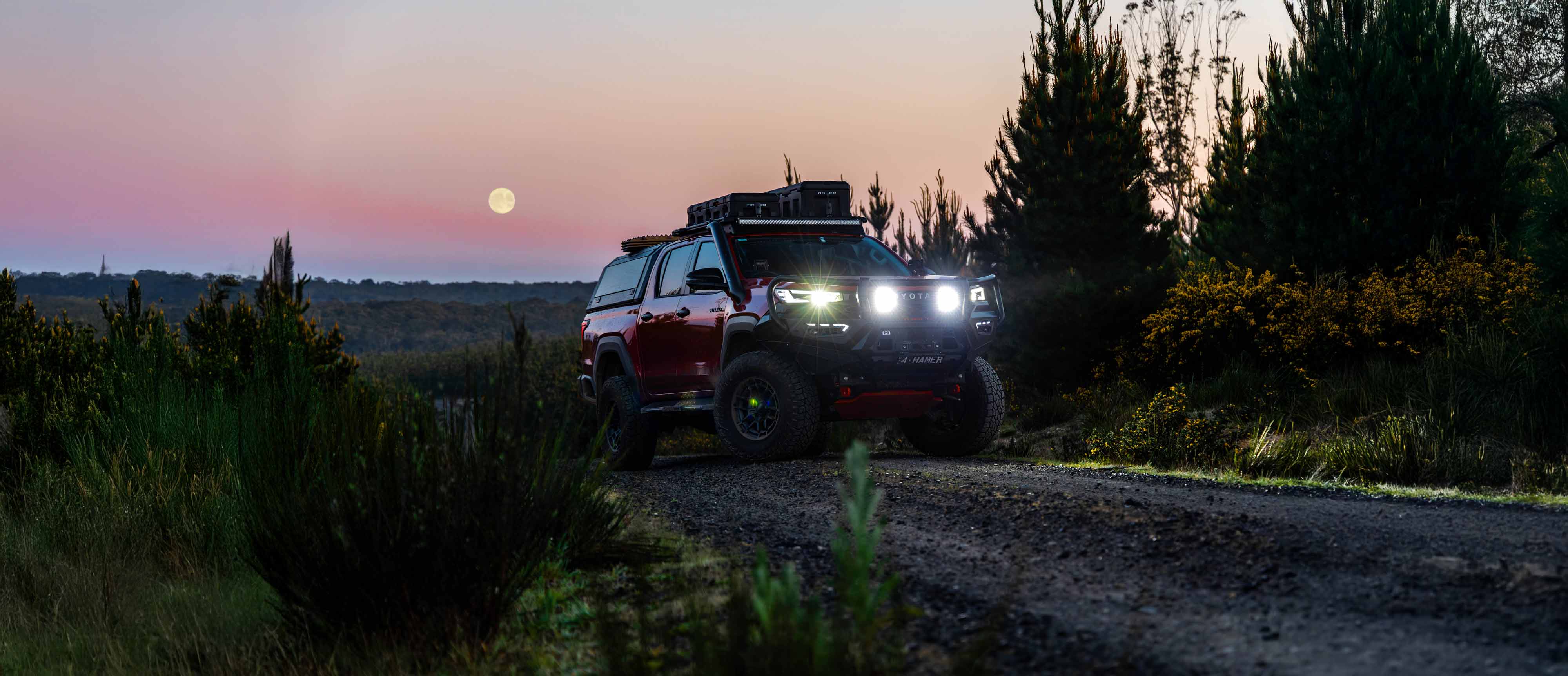 A red  toyota N90 Hilux Ute with roof racks and bright headlights driving on a dirt road at dusk